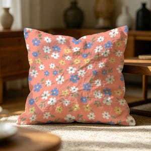 May include: A square throw cushion featuring a coral pink background with a repeating pattern of small white, blue, and yellow flowers interspersed with tiny green leaves. The fabric has a soft, slightly textured appearance with a visible weave.