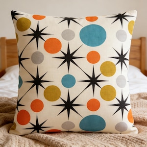 May include: Square decorative pillow with a cream background. The pillow features a repeating pattern of black starbursts and colorful circles in orange, blue, yellow, and gray. The pillow is placed on a bed with a neutral-colored blanket.