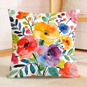 Könnte beinhalten: Ein quadratisches Dekokissen mit weißem Hintergrund und einem lebendigen Aquarell-Blumenmuster. Das Design zeigt große, bunte Blumen in Gelb-, Rot-, Lila- und Orangetönen, akzentuiert durch grüne und dunkelblaue Blätter und Stängel.