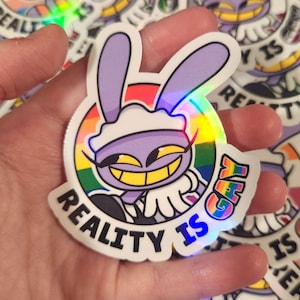 May include: A die-cut holographic sticker featuring a purple cartoon rabbit character with long ears and a wide yellow-toothed grin. The character is set against a circular rainbow background, with the text REALITY IS GAY printed in black.