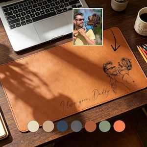 May include: A brown leather desk mat with a sketch of a man and a dog, and the text "I love you, Daddy." A laptop, pencils, a coffee mug, and a notebook are also on the wooden desk. A small photo is in the upper left corner.