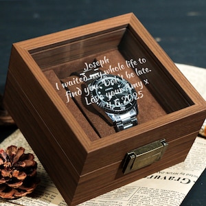 May include: A wooden watch box with a glass top, holding a silver wristwatch with a black dial. The box has a brown interior and a brass clasp. Text on the glass reads: "Joseph, I waited my whole life to find you. Don't be late. Love your Amy x 4.6.2025."