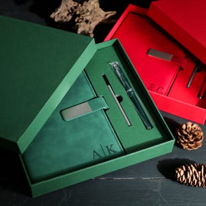May include: A green gift set with a notebook, pen, and card holder. The notebook has the initials "A/K" embossed on the cover. The set is presented in a green box, with a red box visible in the background.