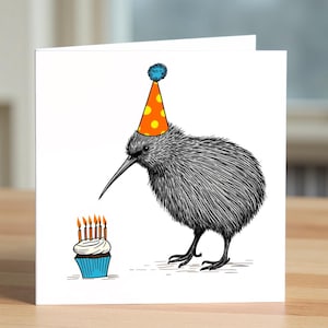 May include: A white birthday card with a black and white drawing of a kiwi bird wearing a party hat with orange spots. The kiwi is next to a cupcake with lit candles. The card is on a wooden surface.