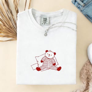 May include: White t-shirt with a red teddy bear graphic wearing striped pajamas and a silver necklace with a pendant. The shirt has a tag that reads "Ongoing Exodos". The shirt is folded and placed on a light-colored surface.