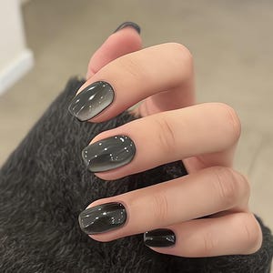 Könnte beinhalten: Eine Hand mit kurzen, oval geformten Nägeln, die in einem Grau-Schwarz-Verlauf lackiert sind. Die Nägel wechseln von einem helleren Grau an der Basis zu einem dunkleren Schwarz an den Spitzen. Die Hand ruht auf einem dunkelgrauen Strickärmel.