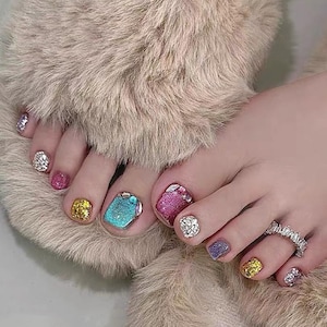 Puede incluir: Primer plano de un pie con una variedad de colores de esmalte de uñas brillantes, incluyendo plata, rosa, oro, turquesa y púrpura. Un anillo plateado con piedras transparentes adorna un dedo del pie. El pie descansa sobre una superficie suave de color marrón claro.