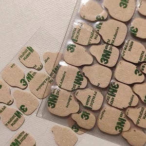 May include: A collection of beige, irregularly shaped adhesive pads with the "3M" logo and text printed in green. The pads are arranged in clear plastic packaging and scattered loosely. The text includes "498PM" and "200QPM Adhesive."