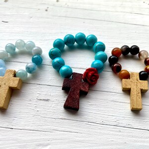 May include: Three wooden cross rosaries with different colored beads. The first rosary has light blue beads, the second has turquoise beads with a red rose charm, and the third has brown and black beads.