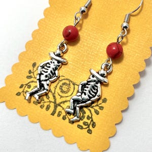 May include: A pair of silver-toned skeleton earrings with red beads. Each earring features a skeleton charm wearing a hat. The earrings are displayed on a yellow, scalloped-edge card with a floral design.