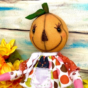 May include: A handmade pumpkin-headed doll with a smiling face, green leaf, and a black cat in a pocket. The doll's outfit features a colorful skirt with orange, brown, and white polka dots. The doll's arms and legs are pink and white.