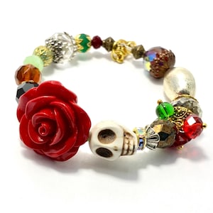 May include: A beaded bracelet featuring a large red rose, a skull, and various colorful beads. The bracelet includes green, brown, and silver beads, with gold accents. The bracelet is a unique accessory.