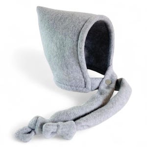 May include: A light gray baby bonnet with a pointed top and a chin strap that fastens with a single button. The bonnet has two long ties that end in small bows. The hat is made of a soft, felt-like material.