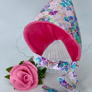 Peut inclure: Un chapeau de fête fantaisiste avec un motif d'écailles de sirène dans les tons de bleu, violet et vert. Le chapeau a un intérieur rose vif et un ruban assorti. Une rose en feutre rose et des feuilles vertes sont à côté du chapeau.