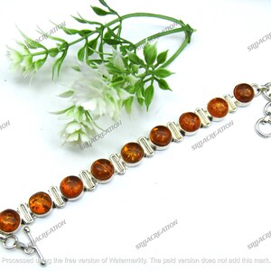May include: A silver bracelet with amber-coloured, round stones. The bracelet has a toggle clasp and is displayed on a white surface with green and white floral accents. The amber stones are evenly spaced along the silver chain.