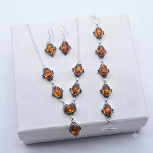 May include: A jewelry set consisting of a necklace, a pair of earrings, and a bracelet. Each piece features oval-shaped, translucent amber stones set in silver-toned metal frames with intricate decorative detailing along the edges.