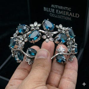 May include: Silver-toned bracelet featuring teardrop-shaped blue gemstones and floral accents. The bracelet is held in a hand, with the text "AUTHENTIC BLUE EMERALD COLLECTION" visible in the background.