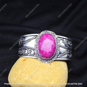 May include: Silver cuff bracelet with a large, oval, bright pink gemstone. The bracelet has an engraved, decorative design with scrollwork and a detailed frame around the gemstone. The jewellery is displayed on a yellow-tan stone against a black background.