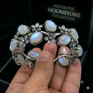 May include: A silver-toned bracelet featuring multiple oval-shaped moonstones with a blue iridescent sheen, interspersed with small metal flower accents. The jewelry is presented in a black box labeled AUTHENTIC MOONSTONE COLLECTION.