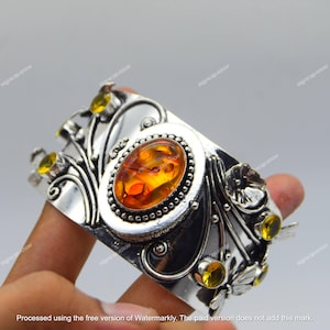 May include: A silver cuff bracelet featuring an oval amber stone at the center, surrounded by a beaded border. The bracelet has floral and leaf designs with yellow accent stones. The bracelet is held by a hand.