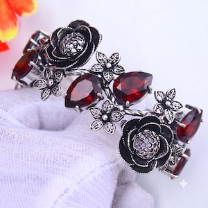 May include: A silver-toned cuff bracelet featuring dark metal floral motifs and faceted deep red teardrop-shaped stones. The design includes detailed metal flowers with textured centres and smaller five-petaled blossoms arranged along the band.