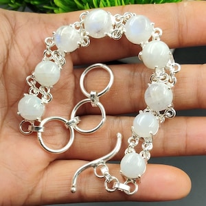 Op de afbeelding: Een zilveren armband met ronde, witte maansteen kralen. De kralen zijn verbonden door een fijne zilveren ketting. De armband heeft een tuimelsluiting. De sieraden worden gepresenteerd tegen een neutrale achtergrond.