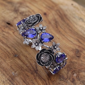 May include: A silver-toned cuff bracelet featuring dark oxidized metal flowers and multiple pear-shaped, deep blue faceted crystals. The design incorporates intricate floral motifs with textured petals and leaves arranged along the open band structure.