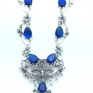 May include: Silver necklace with a butterfly and bird motif, embellished with blue, teardrop-shaped gemstones. The design incorporates floral and leaf elements, offering an elegant, nature-inspired look. The necklace is a statement piece.
