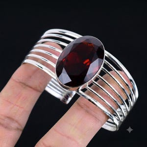 Puede incluir: Un brazalete de plata tipo puño con una gran piedra preciosa de granate rojo oscuro, de forma ovalada y tallada en facetas, engastada en el centro. La banda está formada por siete hilos de plata pulidos, finos y paralelos, con diseño abierto.