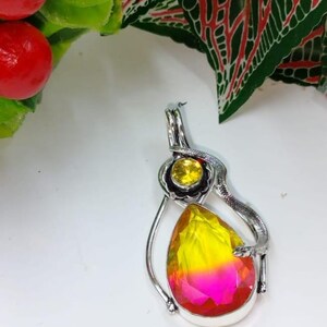 Puede incluir: Colgante de plata con una gran gema en forma de lágrima en tonos amarillo, naranja y rosa. Una piedra amarilla más pequeña se encuentra encima, rodeada por un diseño floral. Un elemento plateado en forma de serpiente envuelve la piedra.