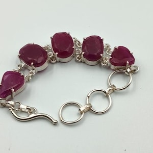 Puede incluir: Una pulsera de plata con piedras preciosas rojas oscuras de forma ovalada y de lágrima. Las piedras están engastadas en engastes individuales de plata y conectadas por eslabones de plata. La pulsera tiene un cierre de broche.