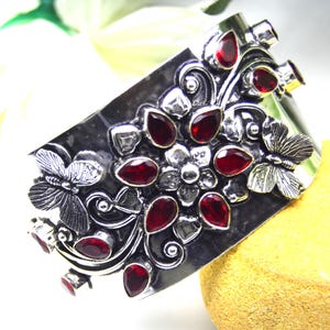 Puede incluir: Un brazalete ancho de plata con un diseño floral y de mariposas. El brazalete presenta varias piedras preciosas rojas oscuras en forma de lágrima. El diseño incluye detalles de plata y se encuentra sobre un fondo brillante.