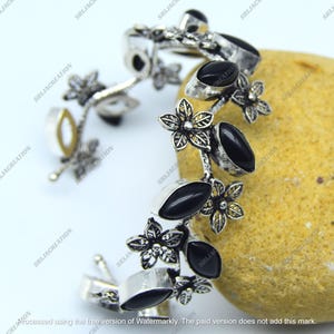 Könnte beinhalten: Silberfarbenes Armband mit schwarzen, ovalen Steinen und floralen Akzenten. Das Armband ist mit kleinen, detaillierten Blüten verziert und vor einem neutralen Hintergrund platziert, um sein filigranes Design zu zeigen.