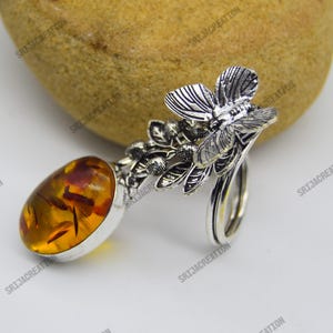 Puede incluir: Anillo de plata con una piedra de ámbar ovalada con inclusiones rojizas. El anillo presenta un diseño floral y de abeja, con intrincados detalles plateados en la banda y los elementos decorativos. El ámbar está engastado en un bisel de plata.
