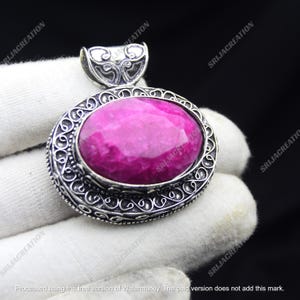 May include: An oval pendant with a large, faceted, magenta gemstone set in a silver-colored filigree frame. The pendant has a decorative bail at the top for a chain. The gemstone is the focal point, with the intricate metalwork adding detail.