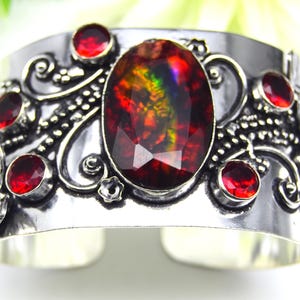 May include: A wide silver cuff bracelet with an oval, multi-coloured gemstone at the centre, surrounded by smaller red gemstones. The bracelet features intricate floral and scrollwork designs.
