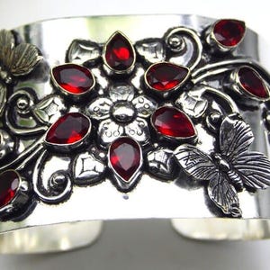 Puede incluir: Pulsera ancha de plata con diseños florales y de mariposas ornamentados. La pulsera presenta piedras preciosas rojas en forma de lágrima. La pulsera mide aproximadamente 5 cm de ancho.