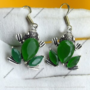 May include: A pair of frog-shaped earrings. Each earring has a green, teardrop-shaped gemstone body, with smaller green leaf accents. The frog's head is detailed with a textured silver finish and a curved hook.