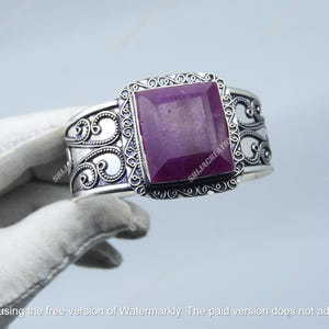 Puede incluir: Un brazalete de plata con un diseño ornamentado y una gran piedra preciosa rectangular púrpura. El brazalete presenta intrincados adornos y un acabado pulido. La piedra preciosa es de un color púrpura intenso.