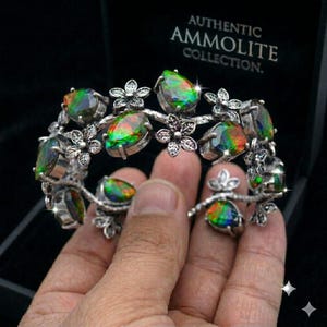 May include: A silver-toned bracelet featuring multiple teardrop-shaped ammolite gemstones with iridescent green and red flashes, interspersed with small metal flower motifs. The bracelet is presented in a black box labelled AUTHENTIC AMMOLITE COLLECTION.