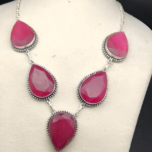 Puede incluir: Un collar plateado con grandes piedras preciosas de color magenta en forma de lágrima. Cada piedra está engastada en un bisel con cuentas y conectada por delicados eslabones. El collar se exhibe en un maniquí blanco.