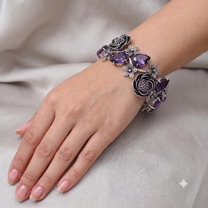 Puede incluir: Una pulsera de tono plateado con piedras preciosas de color púrpura oscuro en forma de pera y motivos florales de metal intrincados. El diseño incorpora pequeños elementos florales texturizados y piedras violetas en un patrón floral continuo.