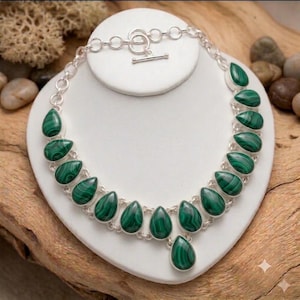May include: A silver chain necklace featuring a series of teardrop-shaped malachite stones with distinct green banding. The necklace is secured with a silver toggle clasp and consists of multiple linked segments, each holding a polished green stone.