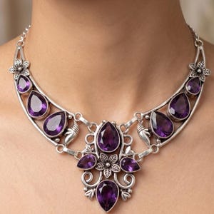 Può includere: Una collana color argento con grandi pietre preziose viola a forma di goccia. La collana presenta un design floreale con pietre viola più piccole e dettagli decorativi argentati. La collana è progettata per essere indossata intorno al collo.