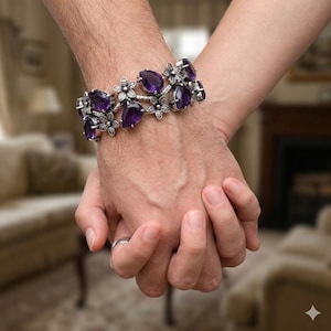 May include: A silver bracelet featuring teardrop-shaped purple gemstones and floral detailing. The bracelet is worn on a wrist, with two hands clasped together. The background is blurred, suggesting an indoor setting.