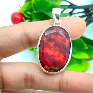 May include: An oval pendant featuring a faceted, deep red gemstone set in a silver-coloured metal. The gemstone has a marbled appearance with darker red and black hues. The pendant is suspended from a silver bail, suitable for a necklace.