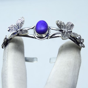 Pode incluir: Pulseira de punho prateada com detalhes de borboletas e uma pedra central ovalada azul escura. O design aberto facilita o uso, e os detalhes de borboleta adicionam um toque de elegância inspirada na natureza a esta joia.