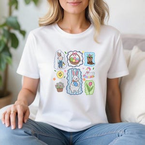 May include: White t-shirt featuring colorful Easter-themed illustrations. The design includes a bunny, Easter eggs, flowers, and other spring motifs. The illustrations are framed in decorative borders, creating a playful and festive look.