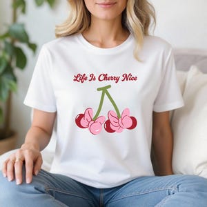 May include: White t-shirt featuring a cherry graphic with the text "Life Is Cherry Nice" in a playful font. The cherries are pink and red with green stems. A casual and fun design.