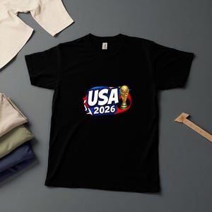 May include: Black t-shirt with a graphic featuring the text "USA 2026" in white, blue, and red, along with a gold trophy. The shirt is made of a soft material.